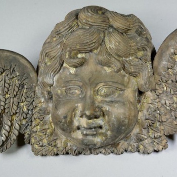Головка ангела с поднятым вверх левым крылом и опущенным правым, почти круглая в лице, с кудрявыми волосами и пухлыми щеками. Позолоченные крылья переданы покрытыми мелкими перьями. Скульптура аналогична с ДС-76.