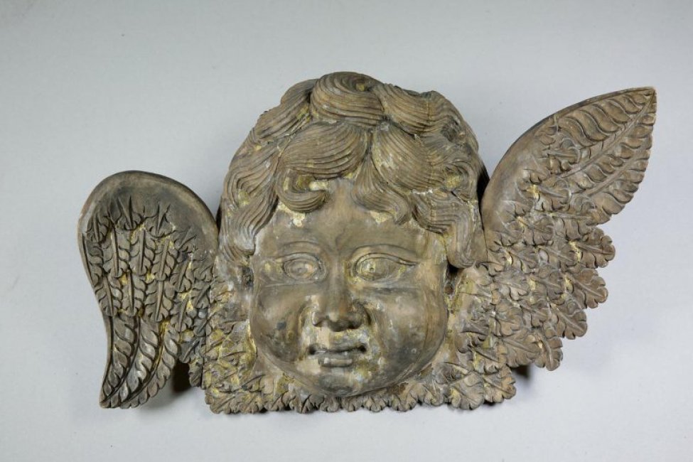 Головка ангела с поднятым вверх левым крылом и опущенным правым, почти круглая в лице, с кудрявыми волосами и пухлыми щеками. Позолоченные крылья переданы покрытыми мелкими перьями. Скульптура аналогична с ДС-76.