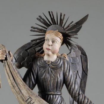 Ангел коленопреклоненный, стоит в нижнем ряду слева. Изображен с поднятой правой рукой и опущенной левой. Одежда посеребрена. Крылья опущенные, посеребренные.