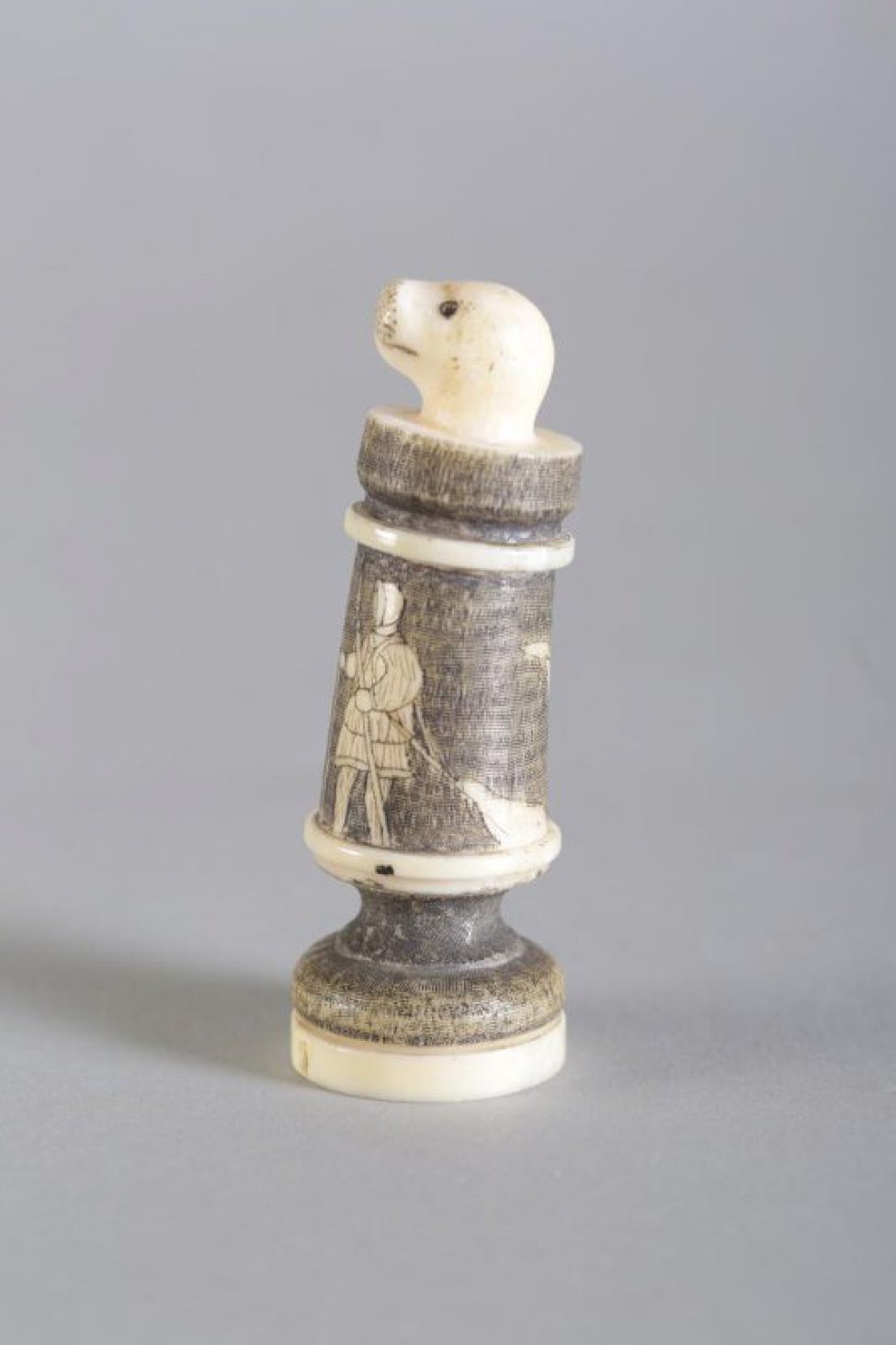 Шахматная фигура в виде цилиндрической профилированной балясины, вверху заканчивается головой нерпы. Голова нерпы  белого цвета, с обозначенными неглубокой резьбой (процарапыванием) маленькими круглыми глазами, линией рта и точками на морде. Средняя  часть покрыта тонированной в черный цвет гравировкой, где изображены в цвете кости две фигуры охотников в зимней одежде с гарпунами в левой руке и с привязанными к плечу убитыми нерпами. Фигуры в 3/4 повороте лицом друг к другу, на обороте шахматной фигуры над нерпами - взлетевшая птица (чайка). Два профилированных пояска и низ постамента белого цвета. Вся фигура выполнена из одного куска бивня.