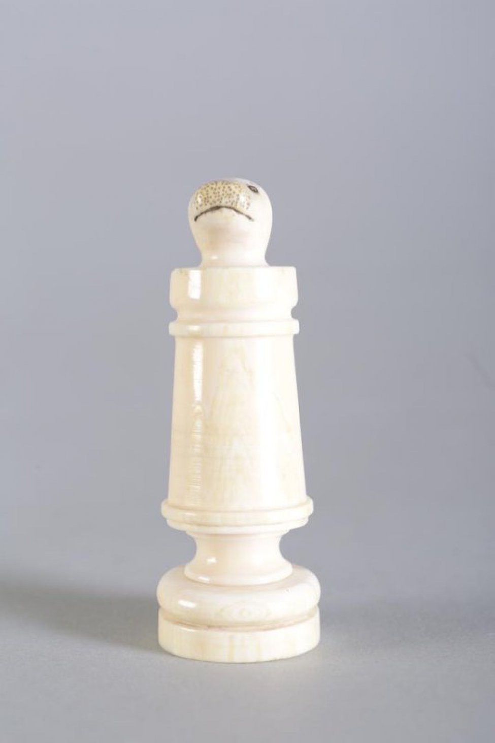 Шахматная фигура белого цвета в виде цилиндрической профилированной балясины, вверху заканчивается головой нерпы. На голове неглубокой резьбой (процарапыванием) с черной тонировкой обозначены  маленькие круглые глаза, линия рта и точки на морде. Вся фигура выполнена из одного куска бивня.