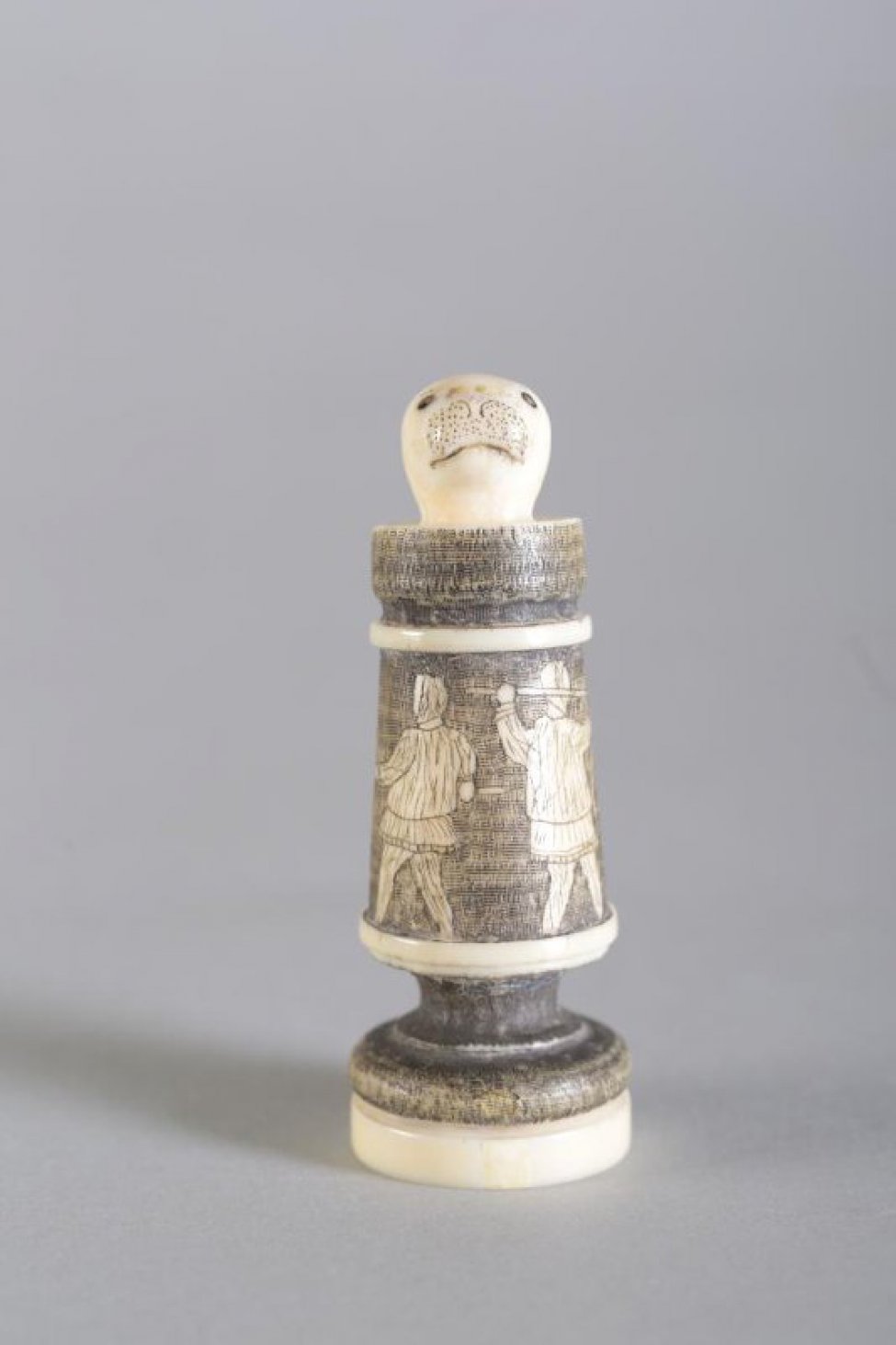 Шахматная фигура в виде цилиндрической профилированной балясины, вверху заканчивается головой нерпы. Голова нерпы  белого цвета, с обозначенными неглубокой резьбой (процарапыванием) маленькими круглыми глазами, линией рта и точками на морде. Средняя  часть покрыта тонированной в черный цвет гравировкой, где изображены в цвете кости две фигуры охотников в зимней одежде, вонзившие с двух сторон гарпуны в огромного моржа с длинными бивнями с повернутой влево головой. Фигуры охотников в 3/4 повороте спинами друг к другу, морж - на обороте  шахматной фигуры. Два профилированных пояска и низ постамента белого цвета. Вся фигура выполнена из одного куска бивня. На дне постамента процарапано «1925 Г.»