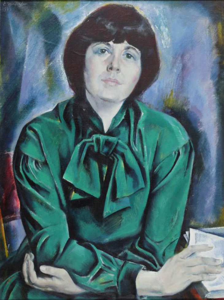 Поясное изображение сидящей за столом темноволосой женщины в зеленом платье с бантом.