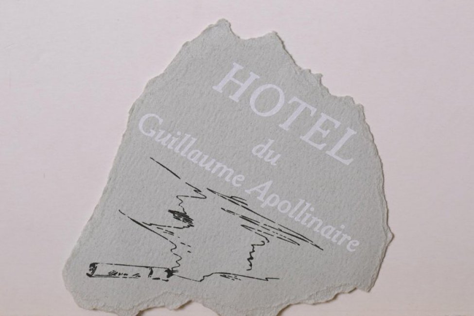 На обрывке серой плотной бумаги изображена дымящаяся папироса, выше - надпись белым цветом на французском языке "HOTEL du Guillaume Apollinaire"