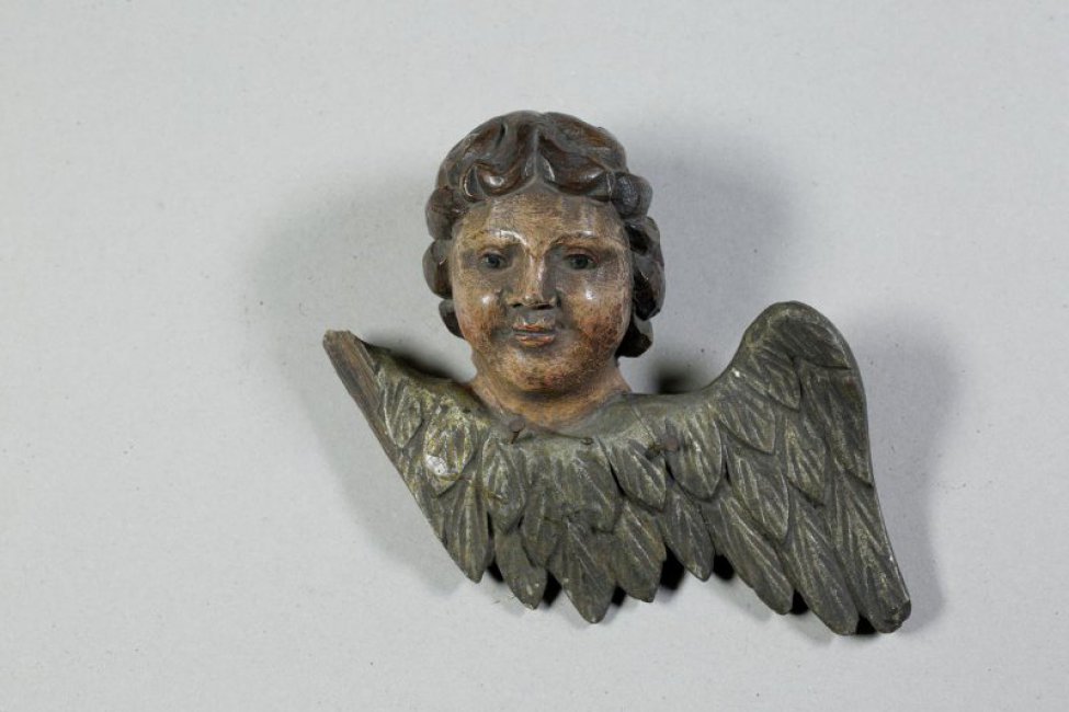 Темнокоричневые волосы на голове ангела изображены в форме прядей, уходящих вниз и назад. Два посеребренных опущенных крыла вырезаны покрытыми 2-мя рядами небольших перьев, рельефно лежащих одни на других.