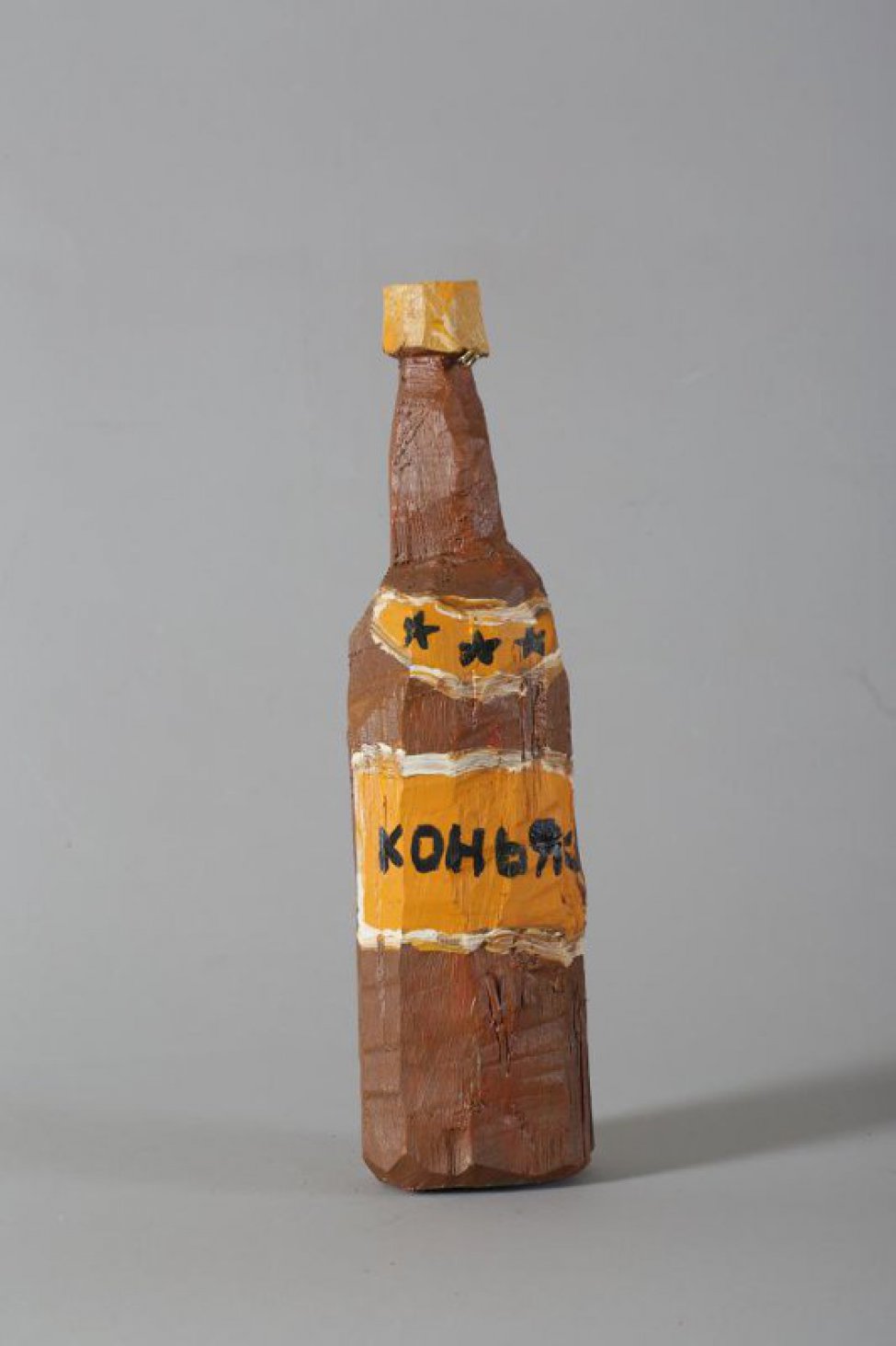 Стилизованное изображение бутылки коричневого цвета, с желтой пробкой. На бутылке 2 желтые этикетки: вверху - полукруглая, с 3 звездочками, внизу - прямоугольная, с надписью КОНЬЯК. Под пробкой металлическое круглое крепление.