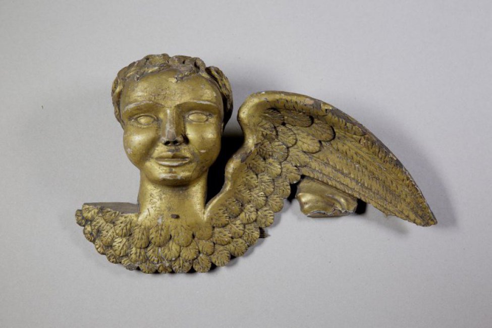 Головка ангела вырезана в резко закругленных поверхностях. Крылья изображены покрытыми 3-мя рядами маленьких перьев. С лицевой стороны скульптура позолочена.