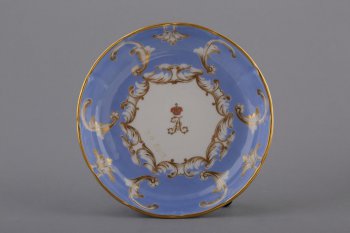 Блюдце чайное голубое, с волнистым золоченым краем, по борту орнамент из белых завитков с золотом; на зеркале в шестиугольном орнаментальном медальоне золоченая буква 