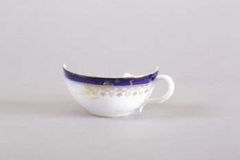 Чашка чайная белая. Тулово с гладкой синей каймой и орнаментальной золоченой каемкой  растительного характера с пятиконечными фигурками по низу каймы.