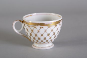 Чашка чайная в форме вазы, белая. По наружной стороне изображена широкая кайма в виде золоченой решетки. По краю снаружи и внутри чашки золоченые каемки.