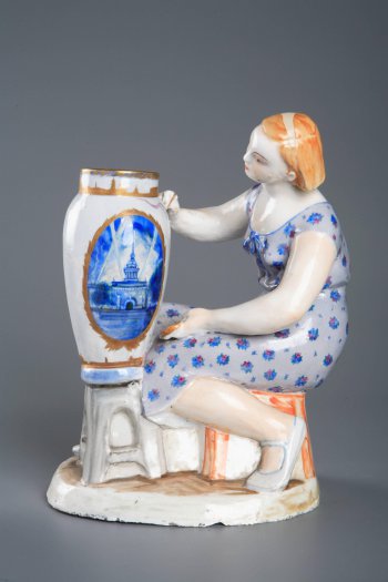 Изображена сидящая молодая женщина в голубом без рукавов платье. Перед ней стоит высокая ваза, которую женщина расписывает.