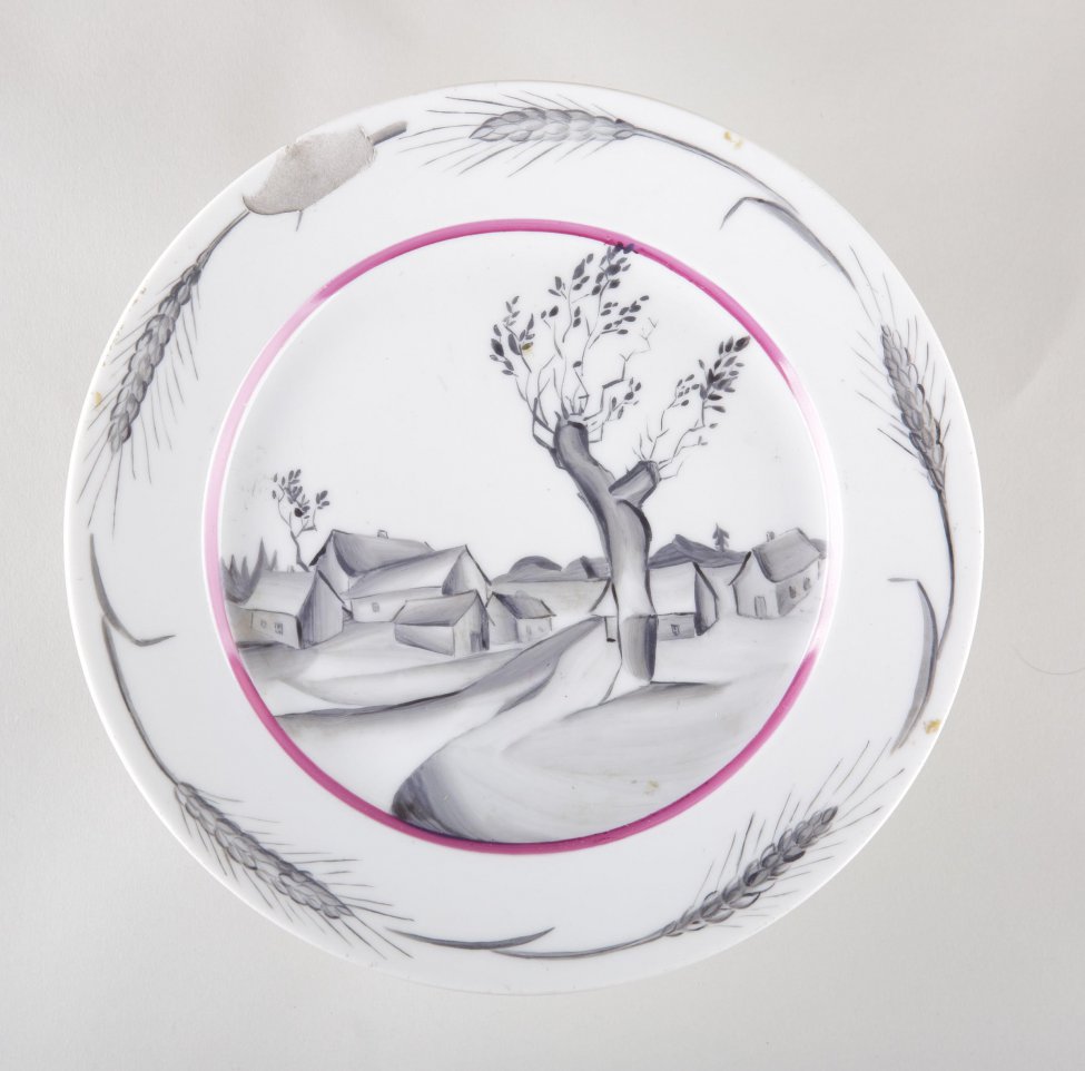 Тарелка мелкая, белая, на зеркале, окаймленном малиновой полосой, изображен серый стилизованный деревенский  пейзаж с домиками и деревом на переднем плане. По борту - пять серых колосьев.