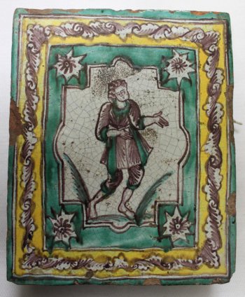 Изразец прямоугольной формы, где на белом фоне коричневым цветом изображена мужская фигура в коричневой куртке с зелеными отворотами и в коротких штанах. Фигура дана в развороте вправо, с согнутыми в локтях и направленными  вправо руками.  Фигура заключена в сложной формы картуш на прямоугольном зеленом фоне с белыми розетками по углам. Вокруг, по краю изразца - желтая прямая рамка с коричневыми стилизованными листьями.