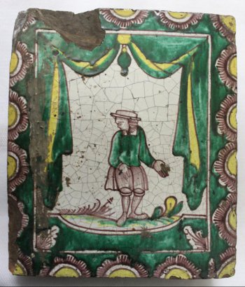Изразец прямоугольной формы, где на белом фоне коричневым цветом изображена мужская фигура, в шляпе, в зеленой куртке и  коротких штанах. Изображен  с разведенными в стороны руками.  Туловище с разворотом вправо, голова - влево. Позем с коричневой травкой, желыми и зелеными листьями.  По внутреннему периметру тонкой рамки дана зеленая с желтым драпировка в виде ламбрекена, низ зеленый. По краям изразца - пояс из половинок цветов с желтыми сердцевинами.