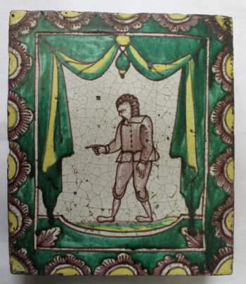 Изразец прямоугольной формы, где на белом фоне коричневым цветом изображена фигура идущего влево мужчины в парике(?), в куртке с пуговицами,  в коротких штанах. Правая рука его с вытянутым указательным пальцем направлена вперед. По внутреннему периметру тонкой рамки дана зеленая с желтым драпировка в виде ламбрекена, низ зеленый. По краям изразца - пояс из половинок цветов с желтыми сердцевинами.