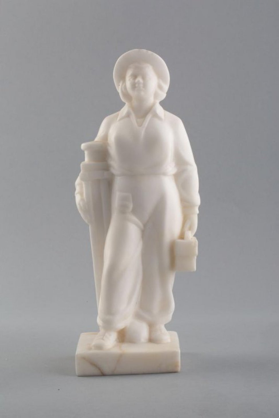 Из белого камня. Изображена женщина в комбинезоне, шляпе, с выставленной вперед ногой. Правой рукой она придерживает штатив, в левой руке держит коробку.