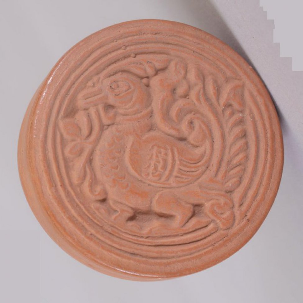 Пресс из розовой глины в форме круга  с плоским верхним и нижним основаниями. Наверху прорезано изображение птицы.