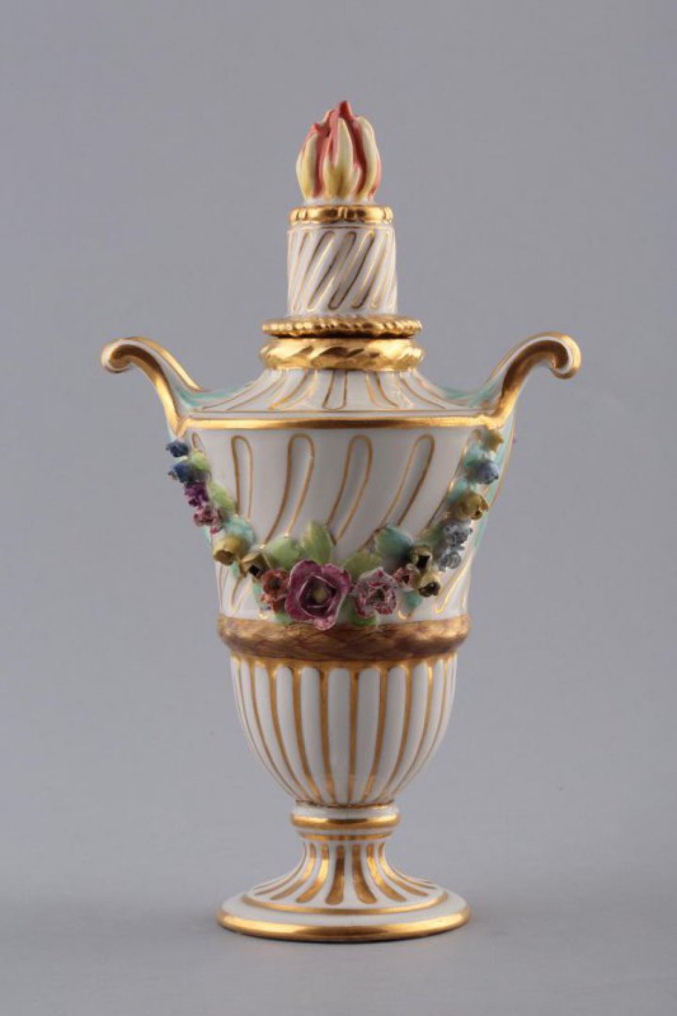 В форме вазы на ножке; с двумя ручками; с переворачивающейся крышкой; по тулову - две гирлянды лепных цветов