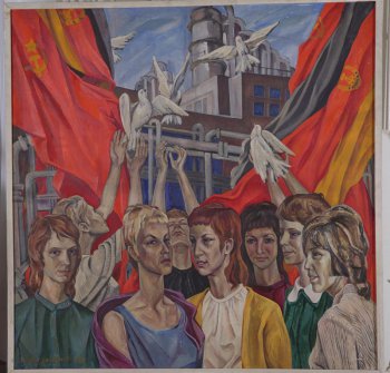 На фоне заводского корпуса и развевающихся слева и справа знамен ГДР и СССР крупным планом погрудно изображена группа молодых женщин (8 чел.). Стоящие сзади пускают в небо белых голубей.