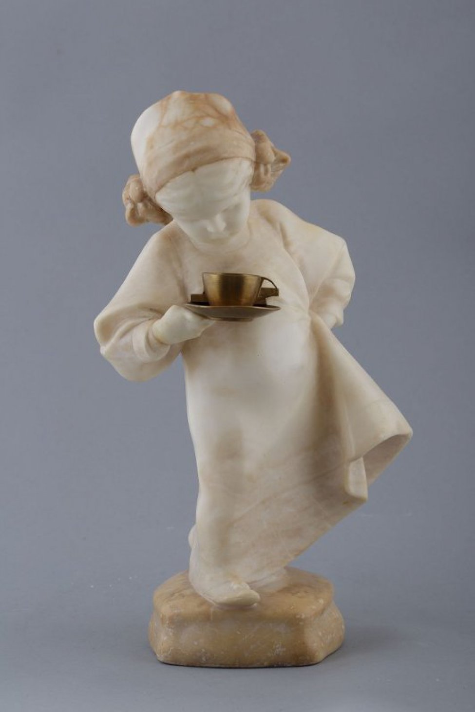 На выпуклом четырехугольном постаменте изображена девочка в широком платье, шапочке с бантиками. В правой руке - чашка с блюдцем, левой рукой придерживает платье, образуя волан. Голова опущена вниз, взгляд устремлен на чашку.