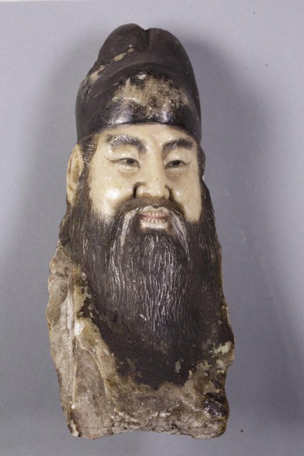 Китайский божок (часть предмета) изображает голову мужчины с китайским типом лица с длинной головой и усами, с полуоткрытым ртом, в котором видны зубы. На голве убор, окрашенный в черный цвет, как и борода, волосы, брови, глаза. Голова не в полном объеме - затылка нет.