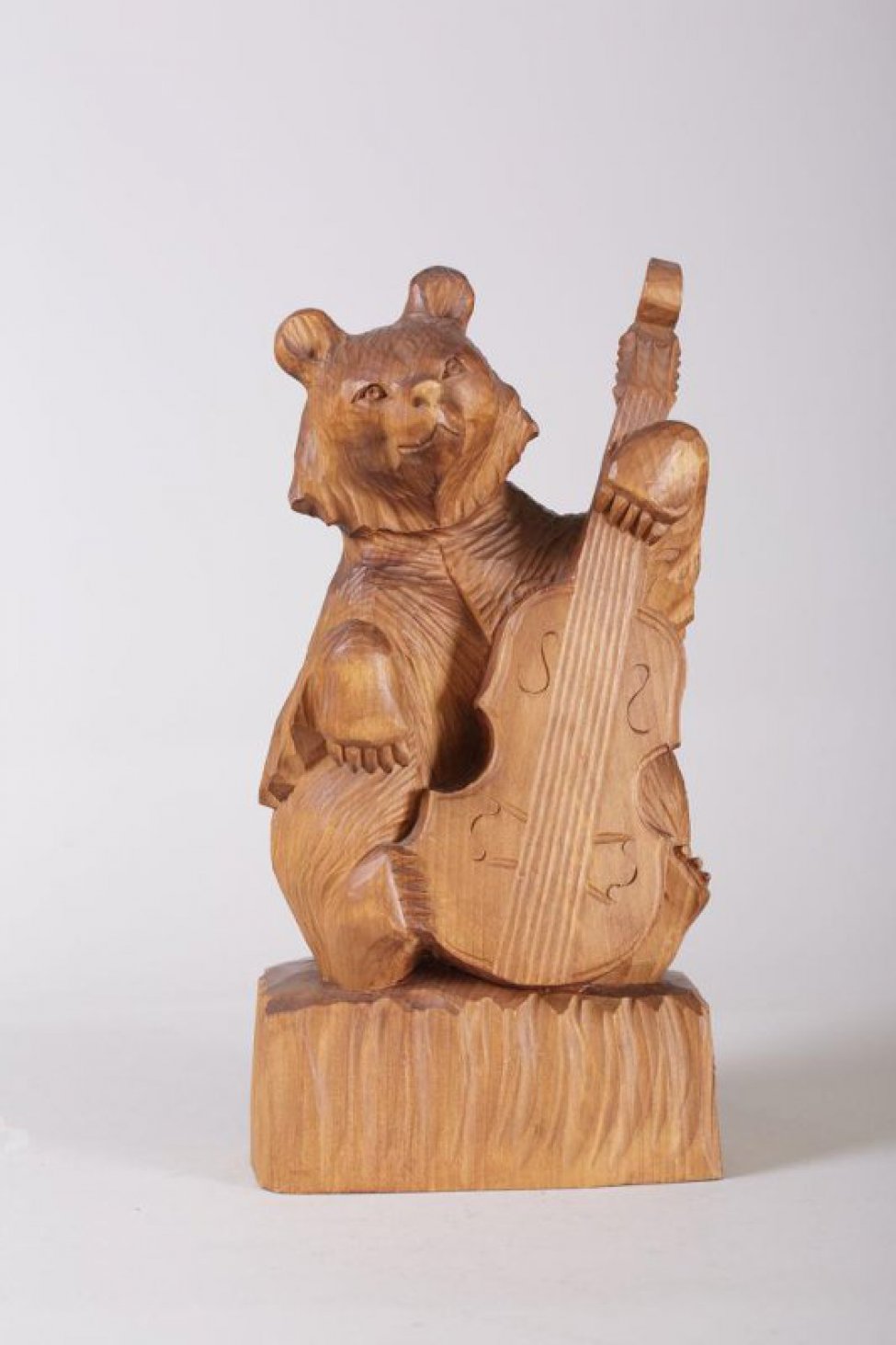 Медведь коричневого цвета сидит, свесив переднюю правую лапу, остальными лапами он держит стоящую перед ним виолончель, подставка семигранная неправильной формы.
