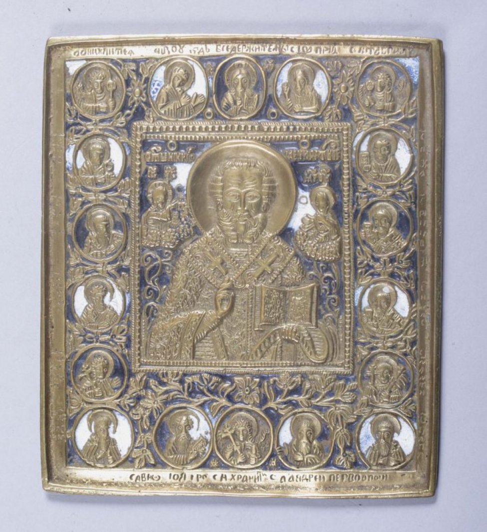 В центре иконы изображен Николай Чудотворец с Иисусом Христом и Марией по бокам. В широкой рамке изображение 18 святых. Фон темно-сининяя и белая эмаль.