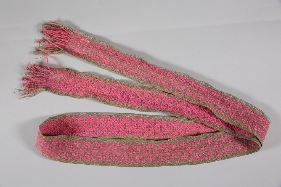 На льняной основе серого цвета и таком же утке розовой шерстью выткан крестообразный узор. Концы украшены каймой (бахромой) из тех же ниток.